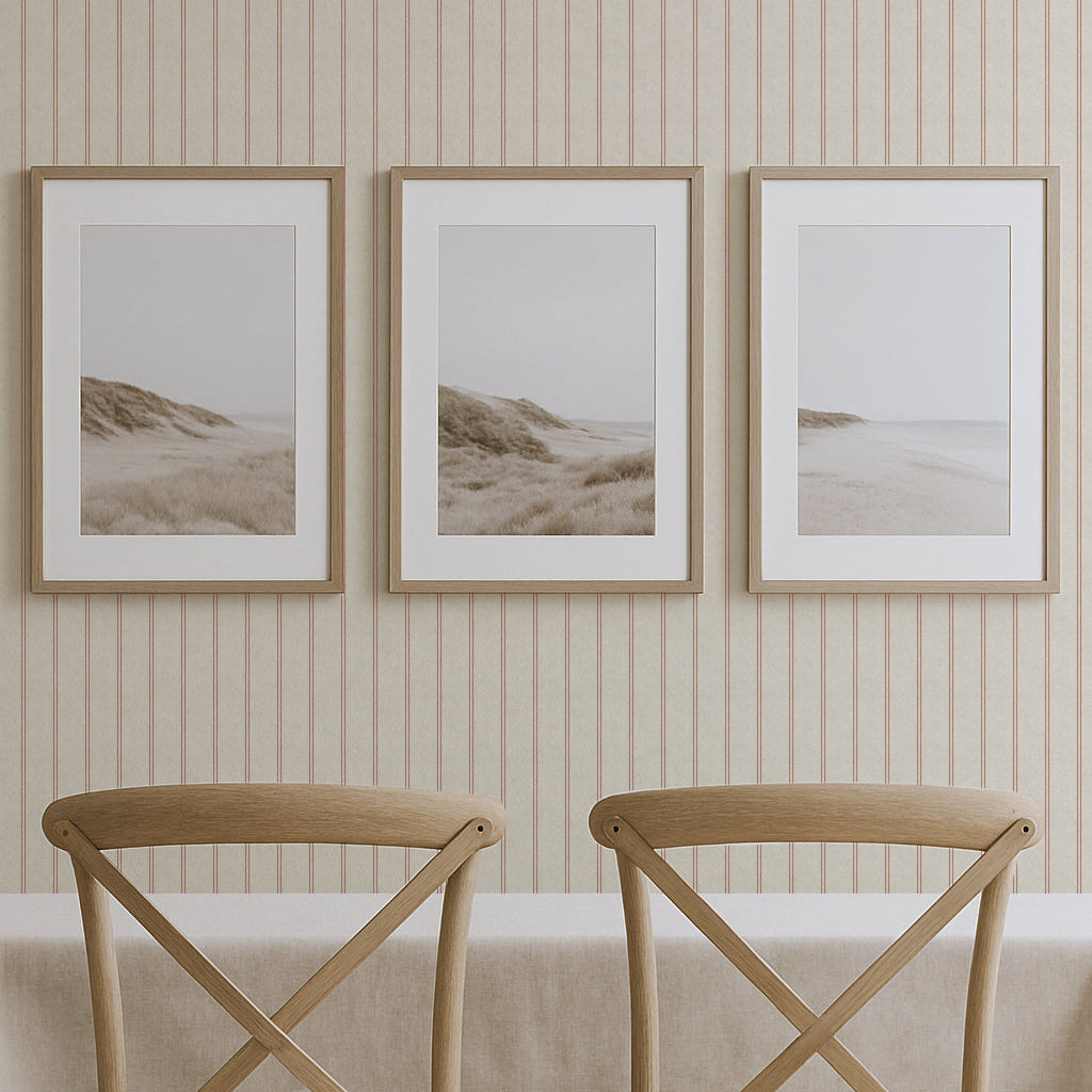 Two wooden chairs with cushioned seats in front of a table with a white tablecloth, against a wall with striped wallpaper and three framed beach scene prints.
